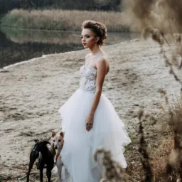 Panna młoda w białej sukni balowej pozuje z dwoma chartami włoskimi na piaszczystej plaży nad jeziorem, z trzcinami w tle.