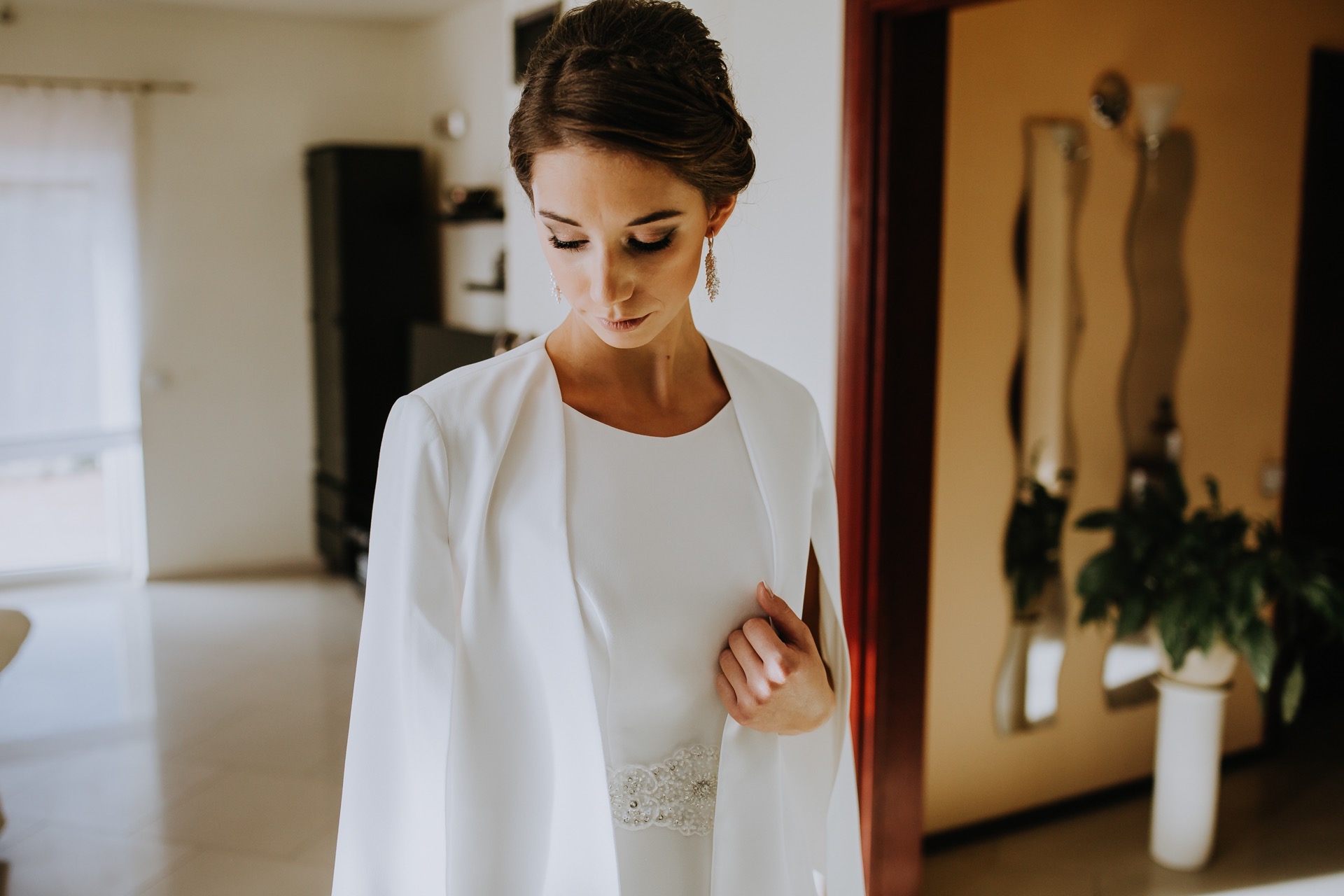 Panna młoda w białej sukni z ozdobnym pasem i narzutką, spoglądająca w dół, z wiszącymi kolczykami.