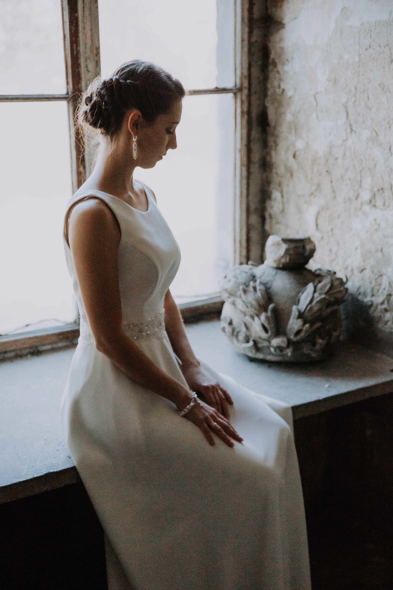 Panna młoda w białej sukni siedzi na parapecie okna w starej kamienicy, z głową pochyloną w dół, obok kamiennej rzeźby.