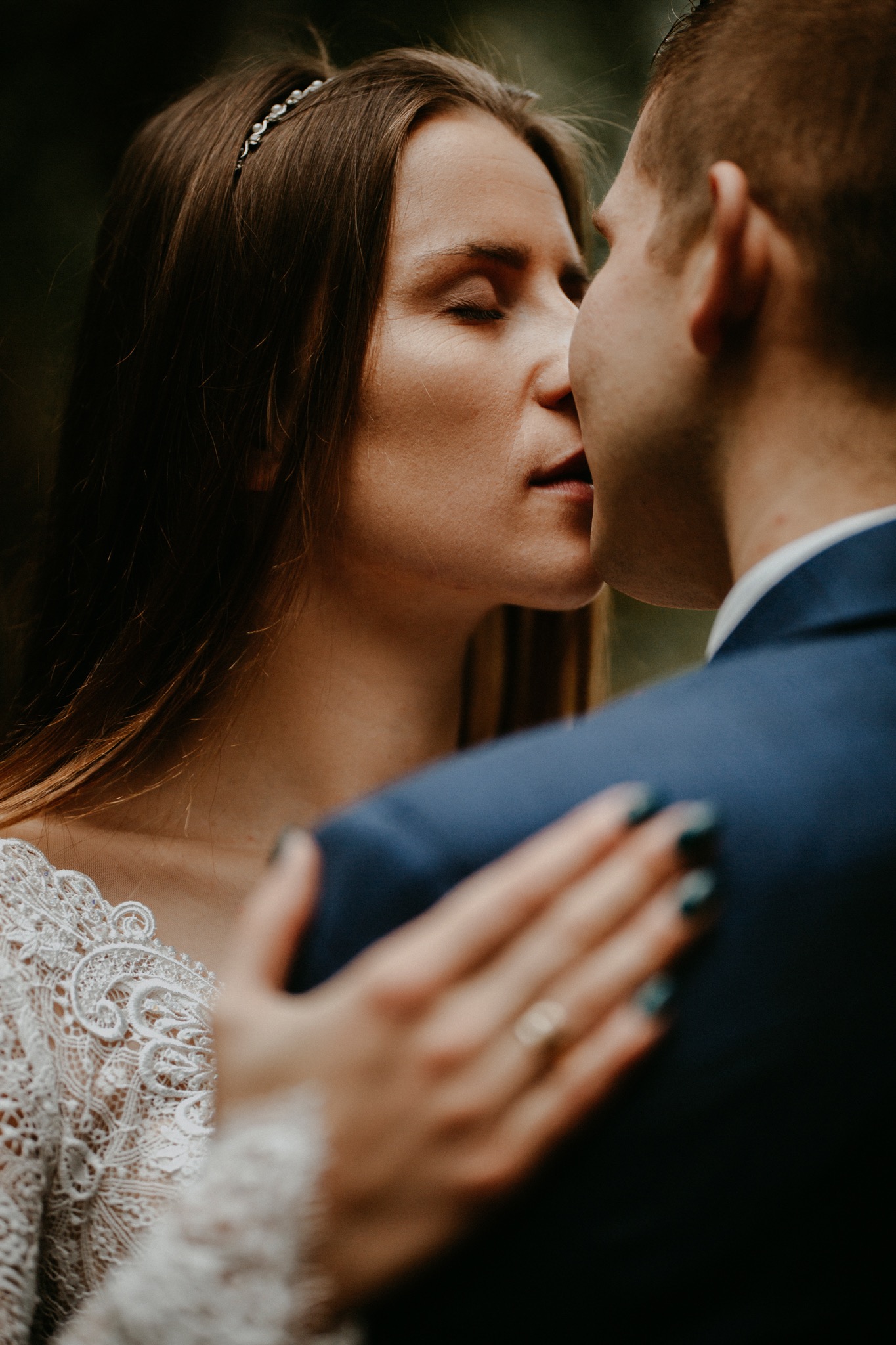 Delikatne zbliżenie na parę młodą w trakcie pocałunku, panna młoda w koronkowej sukni z diademem, pan młody w granatowym garniturze, widoczny pierścionek na palcu panny młodej.