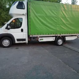 Biały bus z zieloną plandeką, charakterystyczna nadbudowa kabiny, zaparkowany na szarym asfalcie, fragment innego auta w tle.