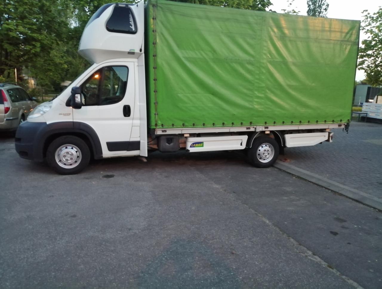 Biały bus z zieloną plandeką, charakterystyczna nadbudowa kabiny, zaparkowany na szarym asfalcie, fragment innego auta w tle.