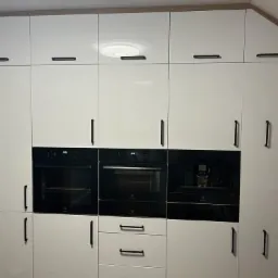 Biała zabudowa kuchenna z piekarnikiem, ekspresem do kawy i szafkami z czarnymi uchwytami, oświetlenie sufitowe LED.