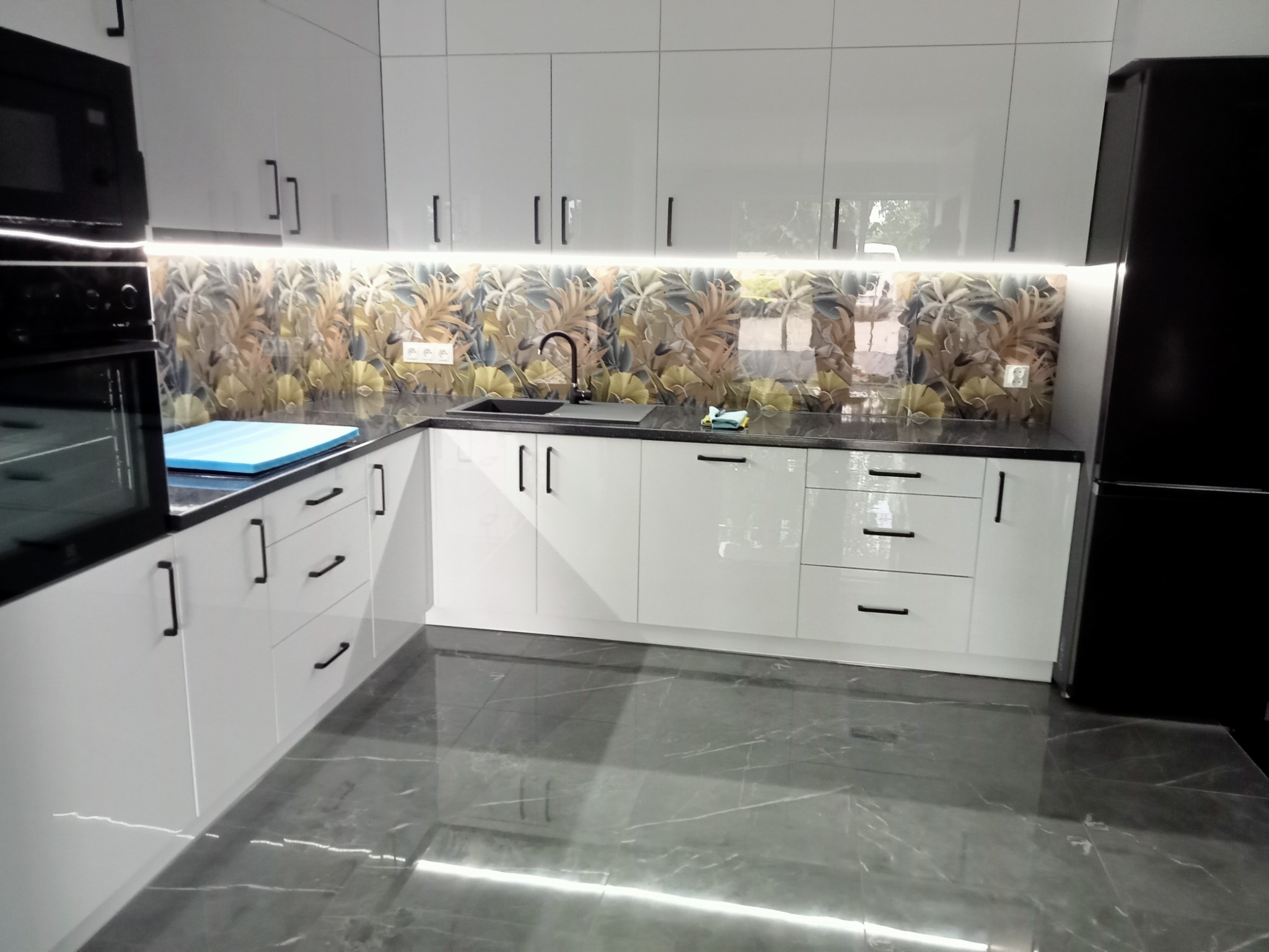 Nowoczesna kuchnia z białymi, lakierowanymi szafkami, czarnym blatem i panelami ściennymi z motywem liści, oświetlona listwą LED pod szafkami wiszącymi, uzupełniona czarną lodówką i szarą podłogą...