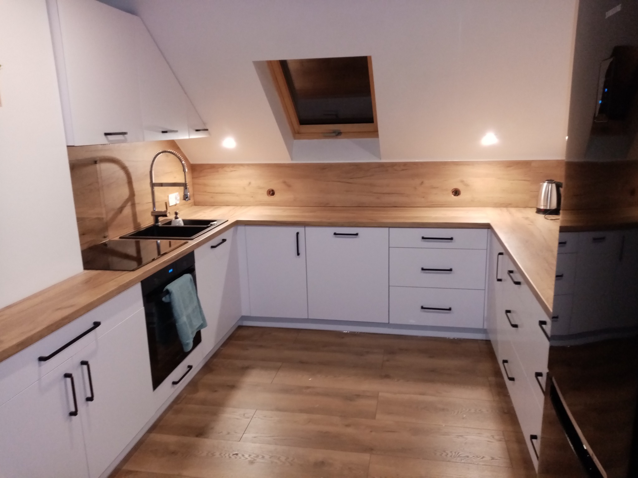 Nowoczesna kuchnia w skandynawskim stylu z białymi szafkami, drewnianymi blatami i oknem dachowym, oświetlona punktowymi lampami.