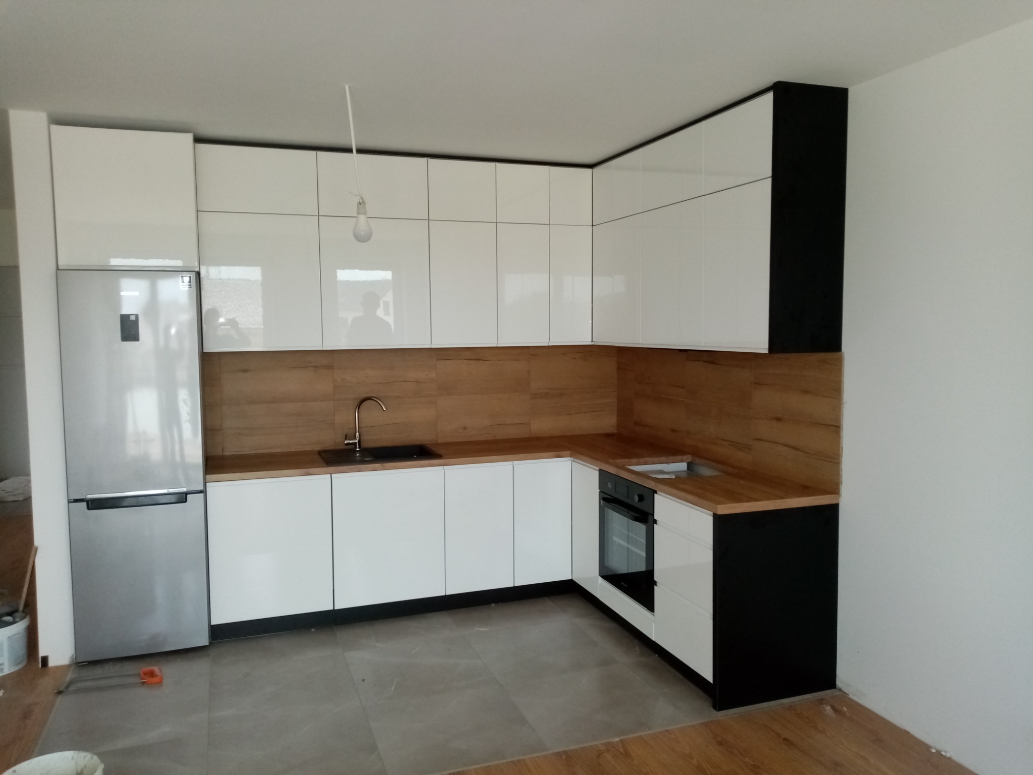 Nowoczesna kuchnia z białymi, błyszczącymi frontami szafek, drewnianym blatem i srebrną lodówką; widoczna surowa żarówka zwisająca z sufitu.