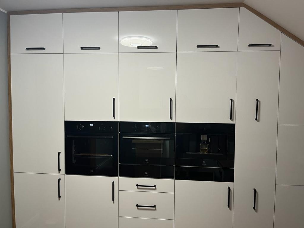 Biała zabudowa kuchenna z piekarnikiem, ekspresem do kawy i szafkami z czarnymi uchwytami, oświetlenie sufitowe LED.