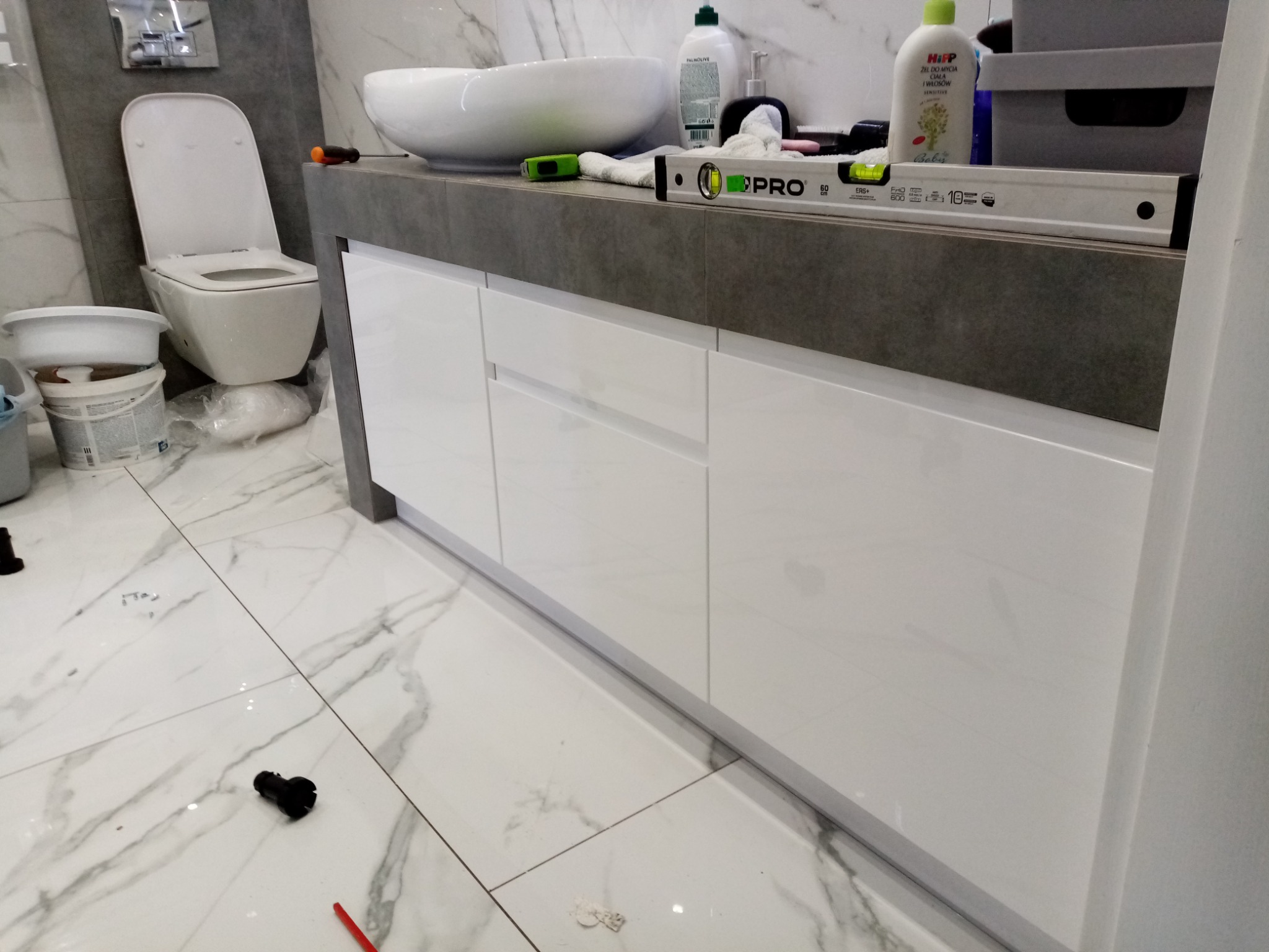Montaż umywalki nablatowej i szafki łazienkowej z białymi, błyszczącymi frontami; widoczny poziomica PRO, narzędzia i akcesoria łazienkowe na blacie oraz toaleta w tle na marmurowej posadzce.