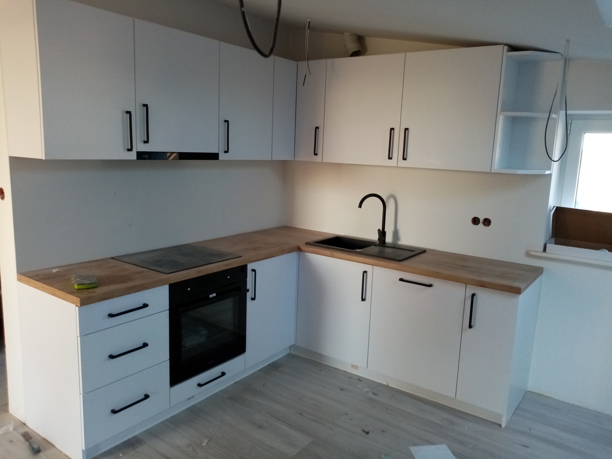 Nowoczesna kuchnia z białymi szafkami i drewnianym blatem, czarnym zlewem i armaturą, płyta grzewcza oraz piekarnik w zabudowie, widoczne niedokończone prace wykończeniowe.