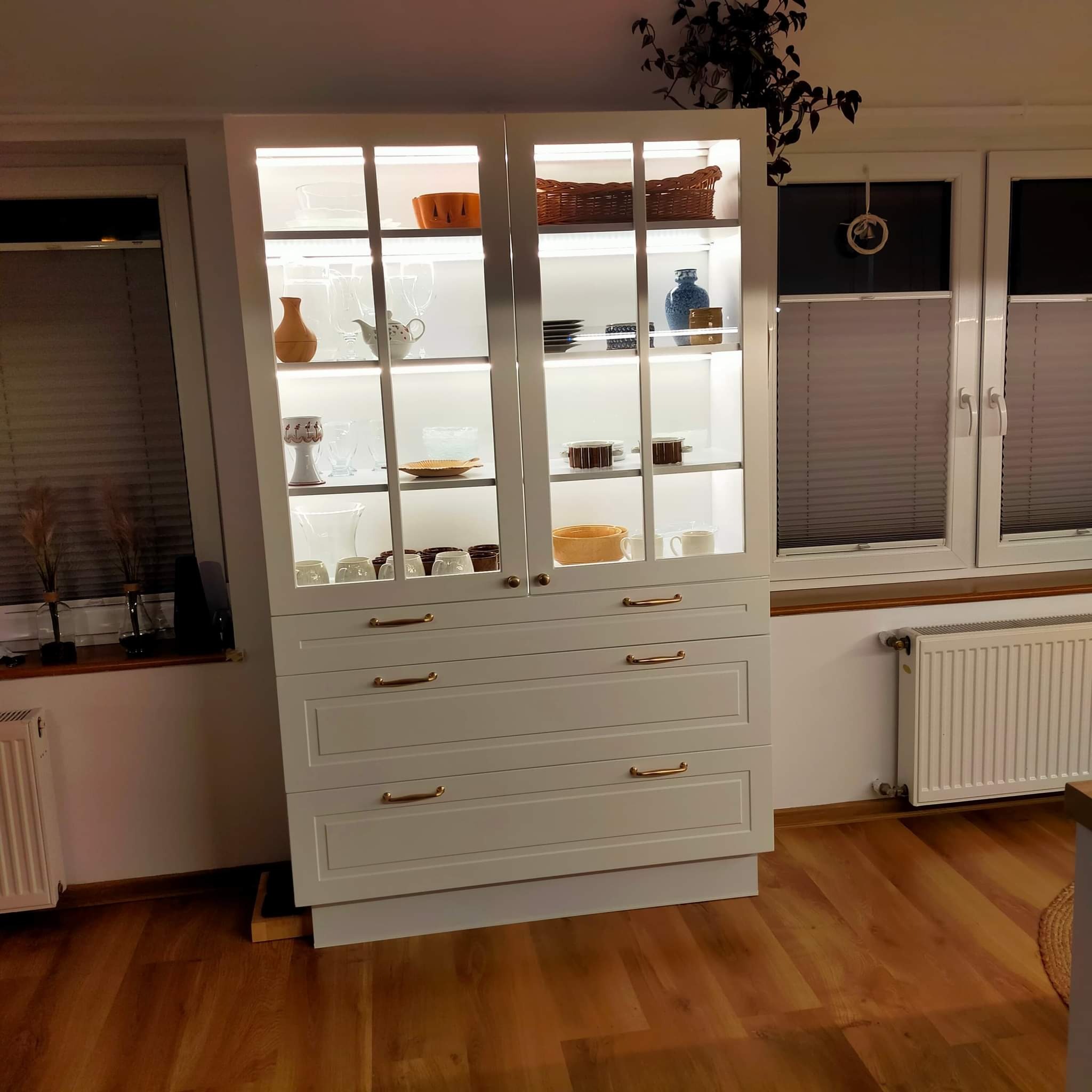 Biały kredens z przeszklonymi drzwiami, podświetlony od wewnątrz, eksponujący kolekcję naczyń i dekoracji, stojący w jasnym wnętrzu obok okna z żaluzjami.