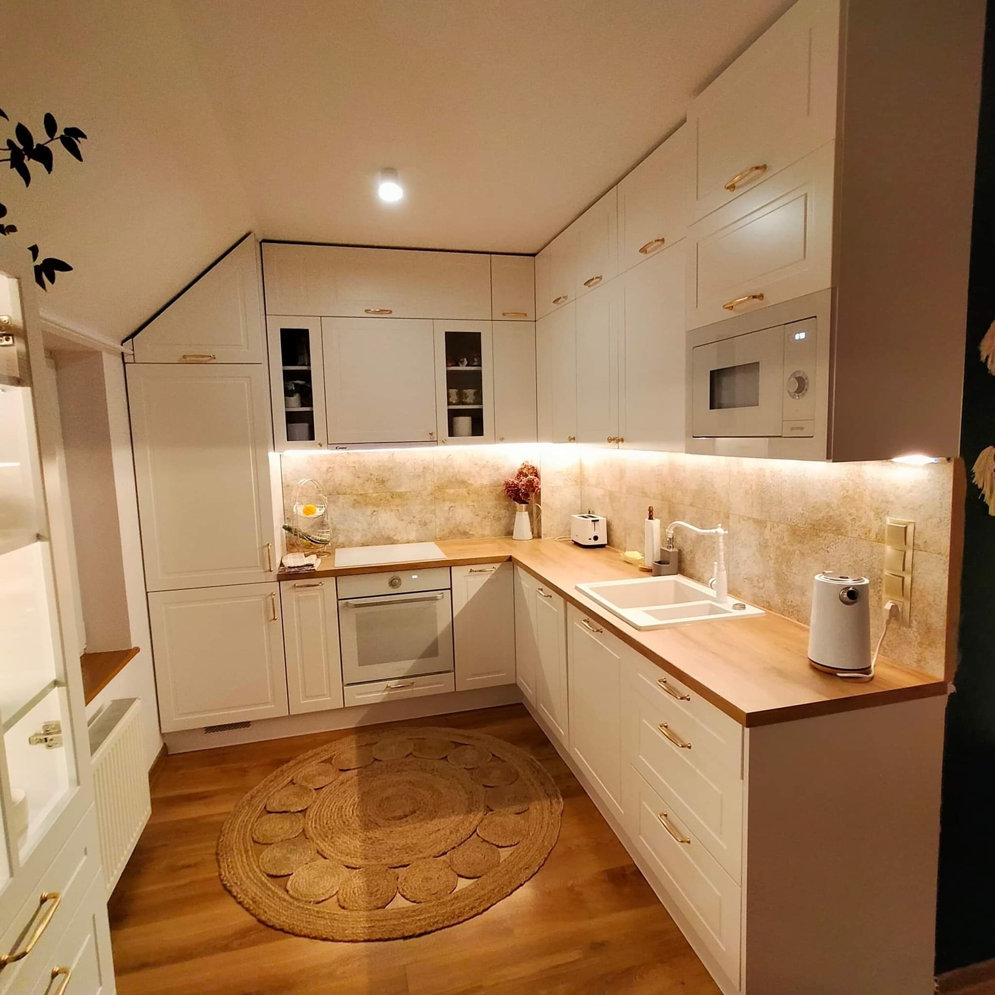 Jasna kuchnia w stylu klasycznym z drewnianym blatem, okrągłym dywanem z juty i dekoracją roślinną na ścianie.