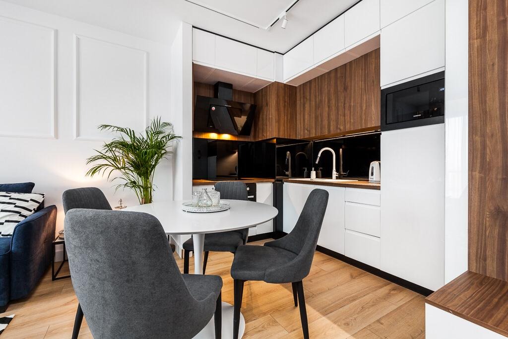 Nowoczesna kuchnia z białymi szafkami i drewnianymi akcentami, okapem nad płytą, okrągłym stołem jadalnym z szarymi krzesłami i rośliną doniczkową.