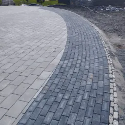 Świeżo ułożona nawierzchnia z szarej i grafitowej kostki brukowej tworząca łuk, obrzeża wykończone jasnymi kamieniami, widoczne przygotowane podłoże i budynki w tle.
