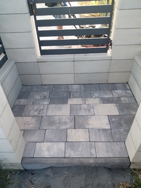 Ułożona kostka brukowa w kolorach szarości tworząca wejście z niskim stopniem, ograniczone słupkami z bloczków betonowych i metalową furtką.