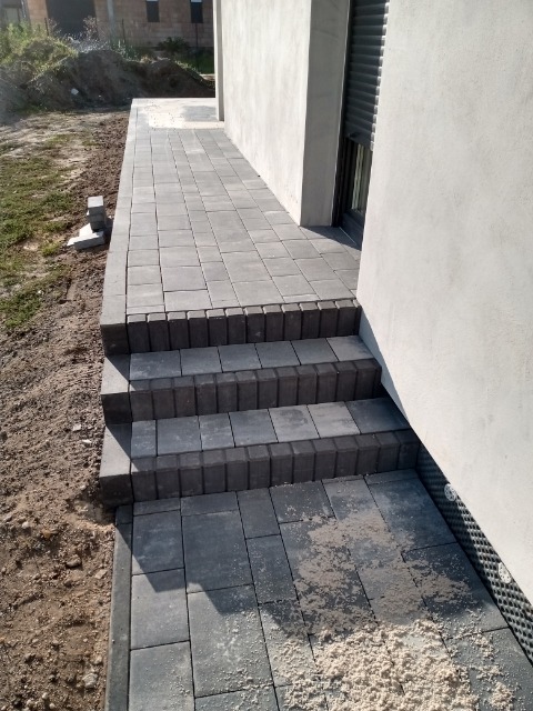 Ułożona szara kostka brukowa tworząca ścieżkę i schody przy budynku z jasną elewacją, widoczne pozostałości piasku na kostce, niezabudowany teren obok ścieżki.