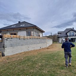 Szwagropol Łukasz Pawlik - Budowa domu jednorodzinnego z bloczków betonowych, widoczna podmurówka z drewnianym szalunkiem, w tle drugi dom i sylwetka mężczyzny na trawniku.