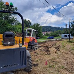 Szwagropol Łukasz Pawlik - Żółty i czerwony minikoparka podczas przygotowywania terenu pod budowę, widoczne wykopane pasy ziemi, tyczki geodezyjne, w tle zabudowa jednorodzinna i zalesione wzgórza.