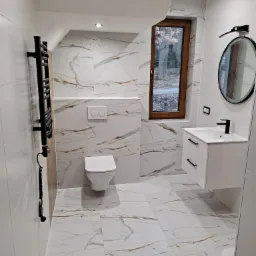 Nowoczesna łazienka z białymi płytkami imitującymi marmur, czarnym grzejnikiem drabinkowym z kablem, wiszącą toaletą i szafką pod umywalkę z czarną armaturą, okrągłe lustro w czarnej ramie...