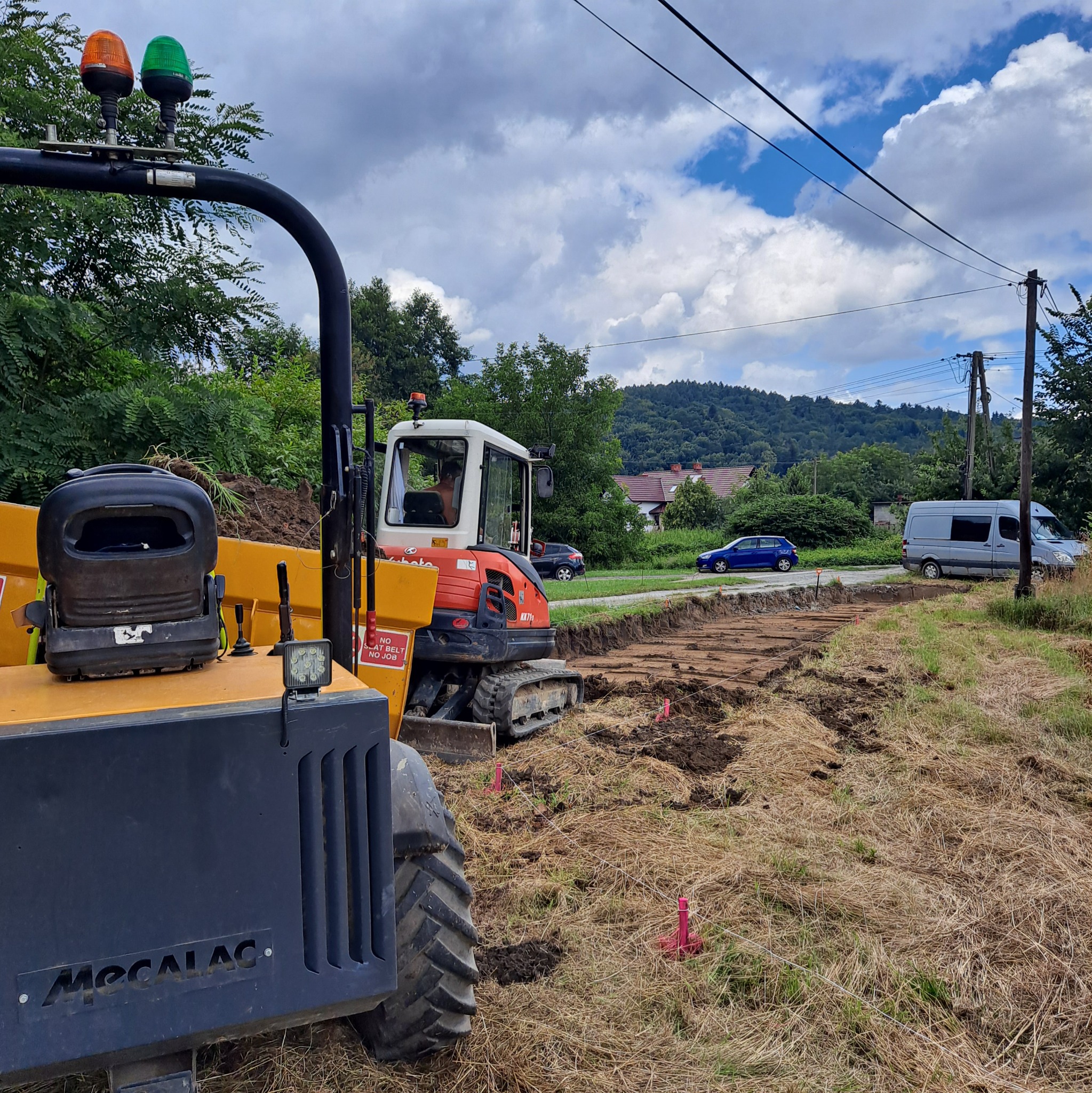 Żółty i czerwony minikoparka podczas przygotowywania terenu pod budowę, widoczne wykopane pasy ziemi, tyczki geodezyjne, w tle zabudowa jednorodzinna i zalesione wzgórza.