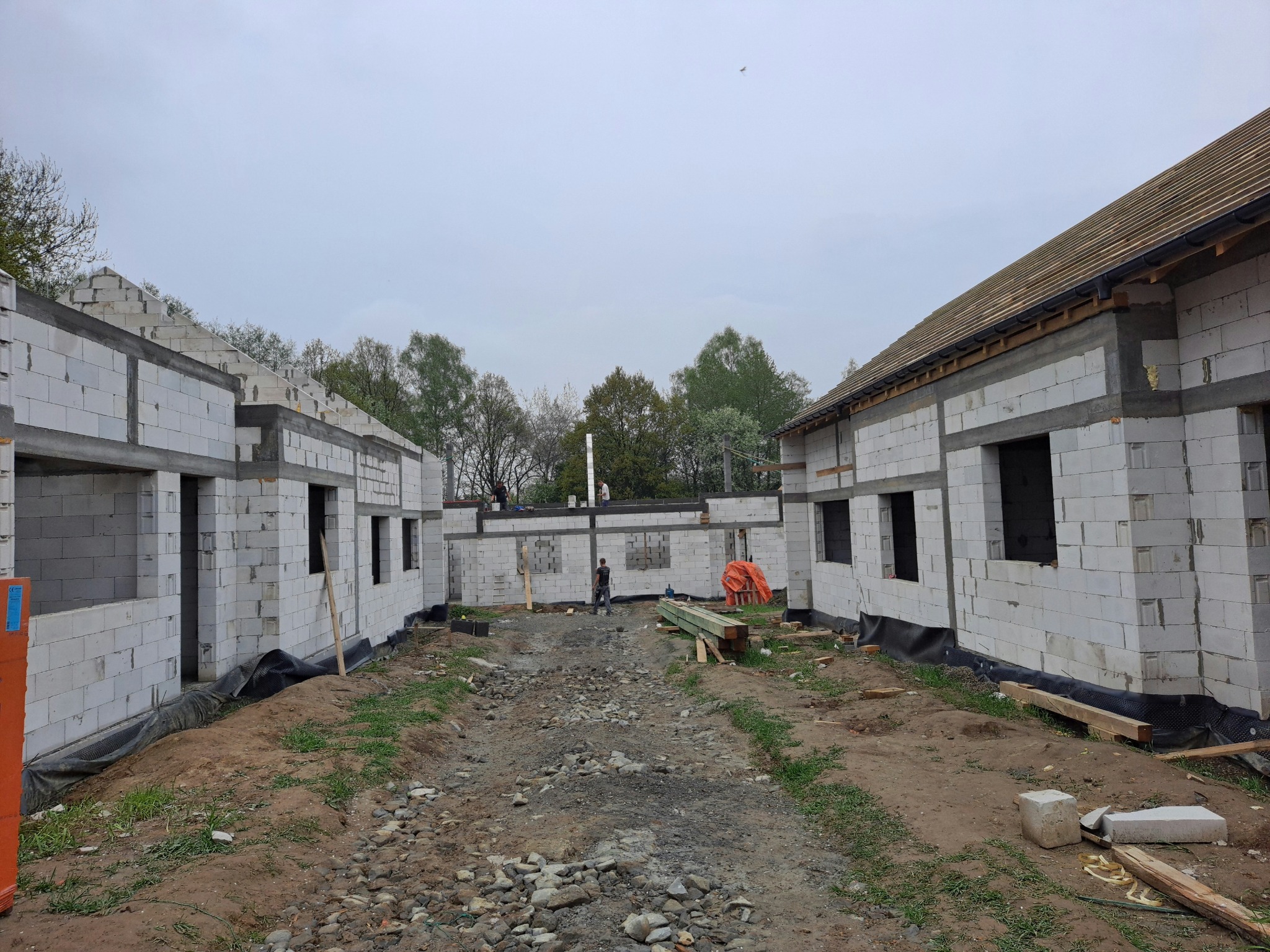 Budowa dwóch domów jednorodzinnych z białych bloczków, widoczne ściany nośne i przygotowane otwory okienne, teren budowy z gruzem i materiałami budowlanymi, w tle drzewa pod pochmurnym niebem.