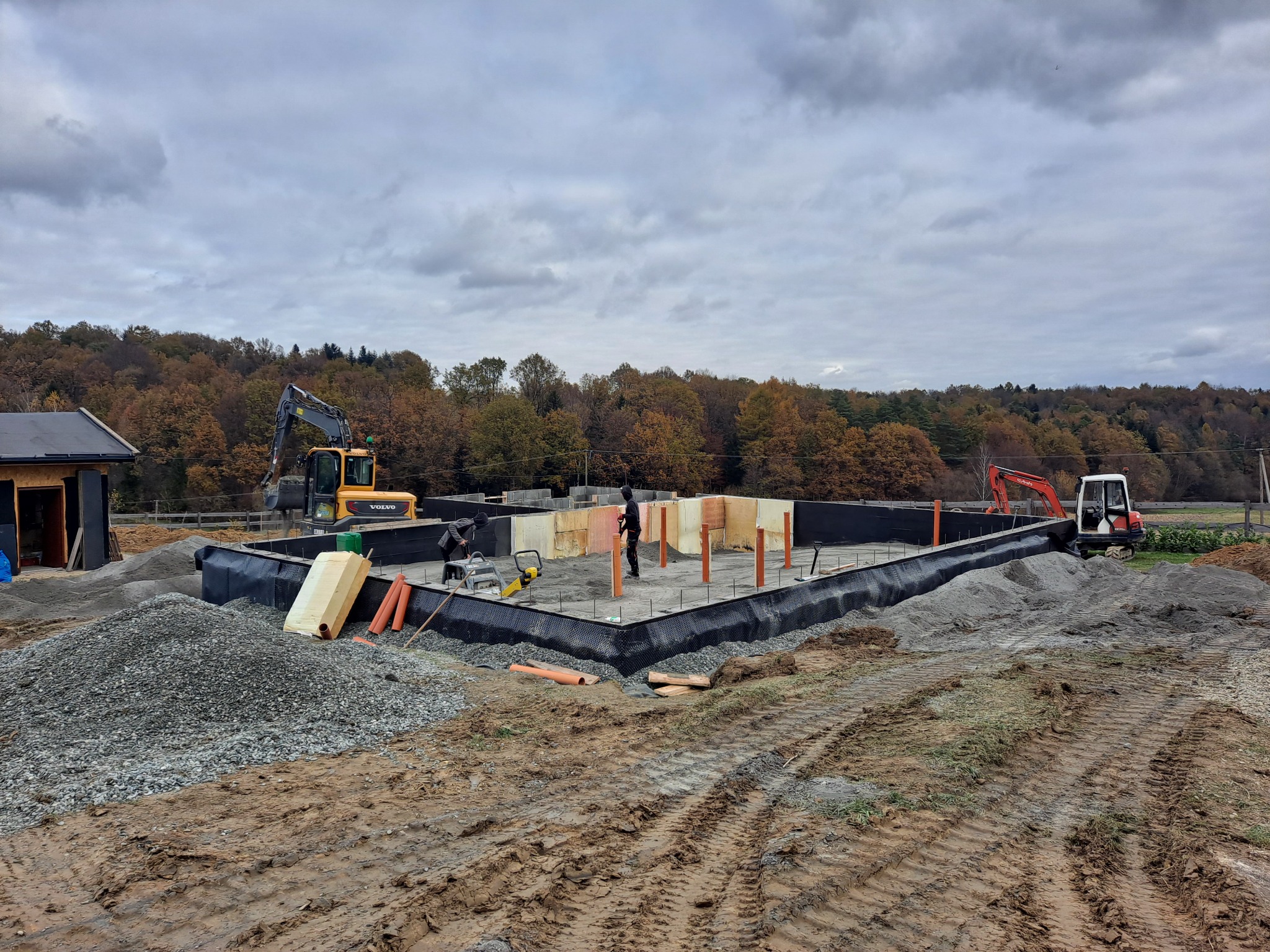 Budowa fundamentów domu: wylewka otoczona czarną folią, pomarańczowe rury, żółta koparka Volvo w tle, jesienny krajobraz.
