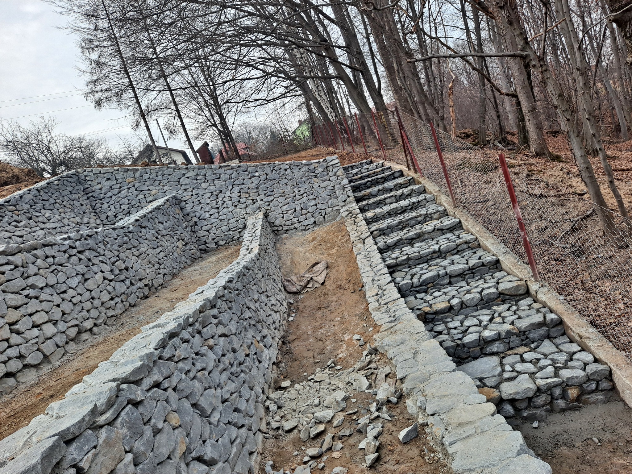 Kamienne schody i murek oporowy w trakcie budowy na zboczu, widoczne drzewa w tle.