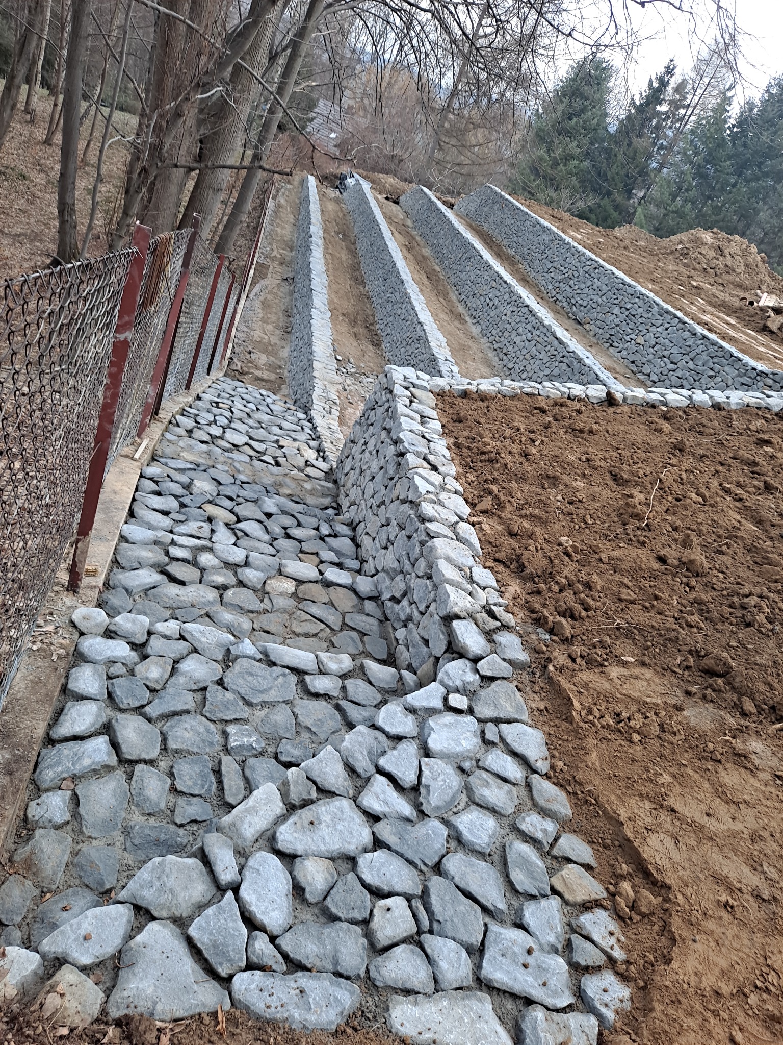 Skarpa wzmocniona kamiennymi gabionami i ścieżką z kamienia naturalnego, zabezpieczona siatką ogrodzeniową po lewej stronie.
