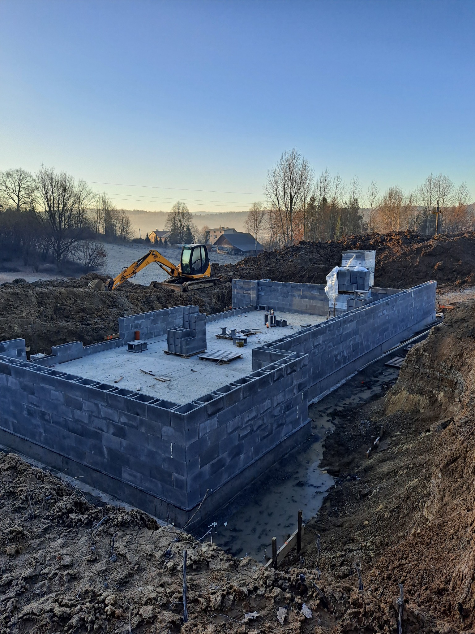 Fundamenty domu z bloczków betonowych w trakcie budowy, częściowo zalane wodą, z koparką w tle i krajobrazem wiejskim w porannym świetle.