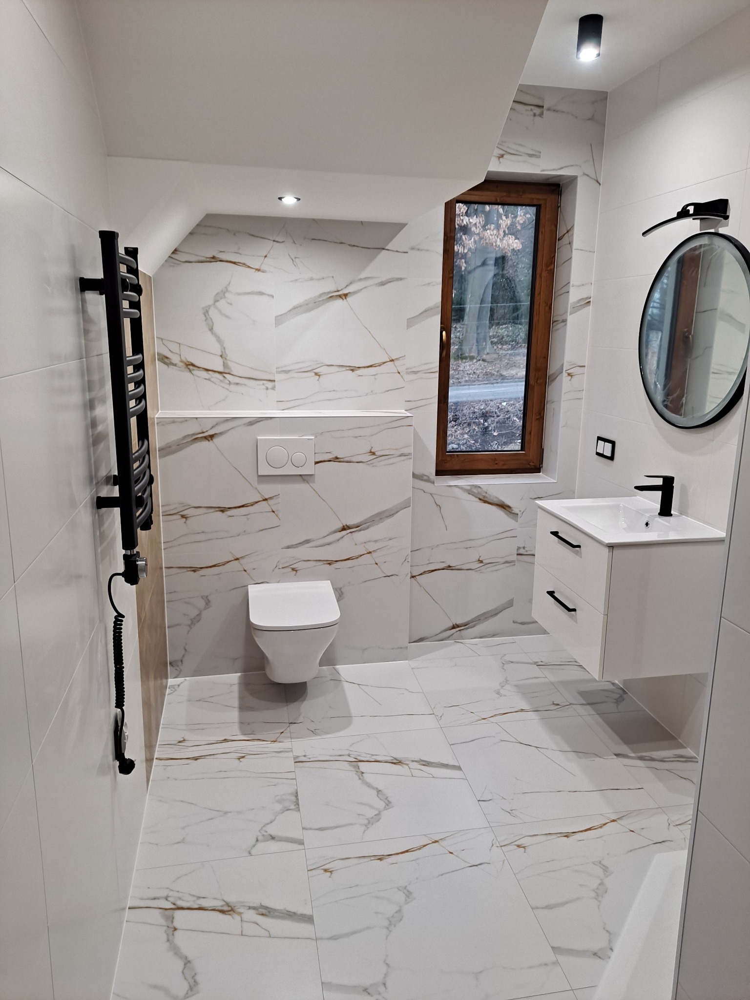 Nowoczesna łazienka z białymi płytkami imitującymi marmur, czarnym grzejnikiem drabinkowym z kablem, wiszącą toaletą i szafką pod umywalkę z czarną armaturą, okrągłe lustro w czarnej ramie...