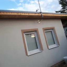 Elewacja budynku z dwoma oknami, wykończona jasnym tynkiem strukturalnym i brązowymi obramowaniami okiennymi, widoczna antena satelitarna na dachu i wiadro z farbą przy ziemi.