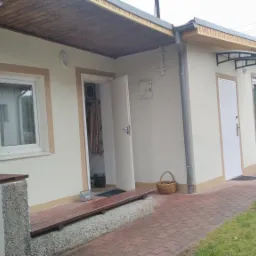 Elewacja domu jednorodzinnego po renowacji, z widocznymi oknami, drzwiami wejściowymi i wykończonym cokołem. Na zewnątrz koszyk wiklinowy i mata pod drzwiami.