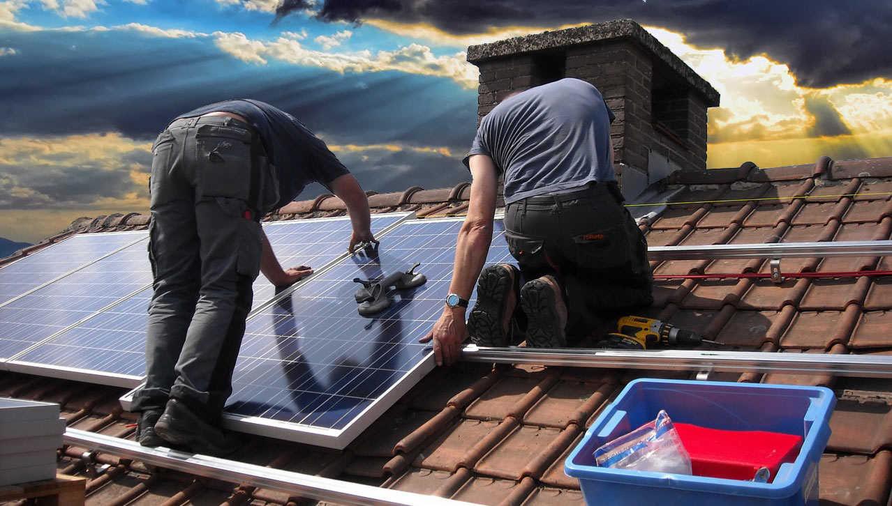 Montaż paneli słonecznych na dachu z cegły, dwóch instalatorów w roboczych ubraniach, narzędzia w niebieskim pojemniku, widok na pochmurne niebo z promieniami słońca.