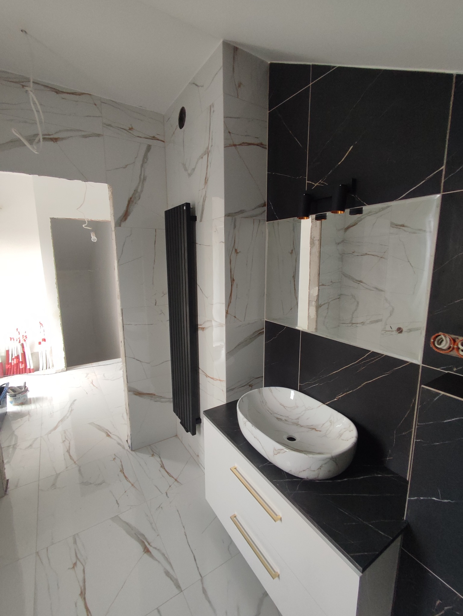 Łazienka w trakcie wykańczania: biało-czarne płytki imitujące marmur, owalna umywalka nablatowa na białej szafce z złotymi uchwytami, pionowy grzejnik panelowy, widoczne niedokończone instalacje.