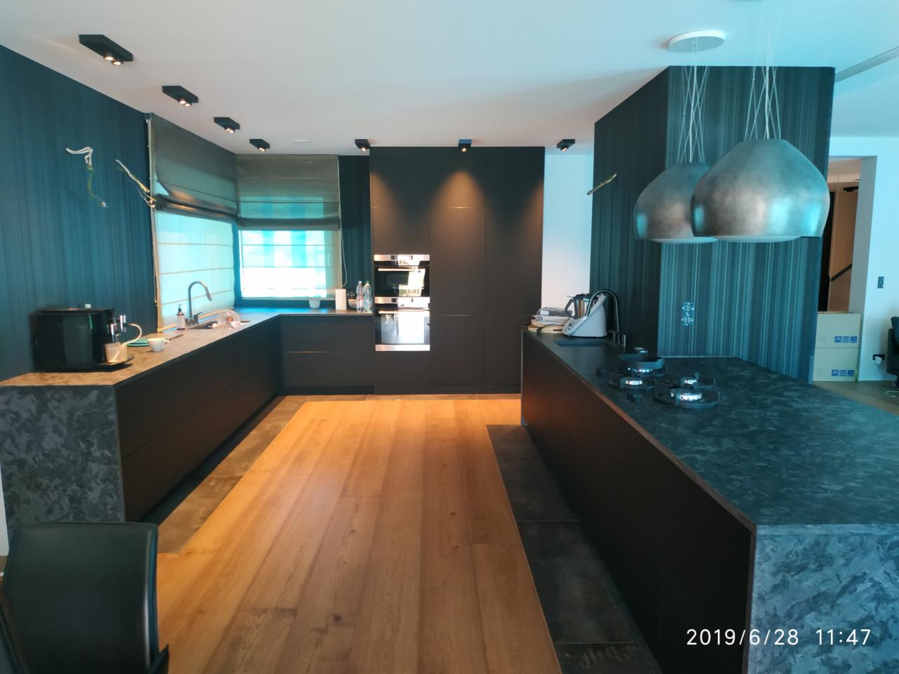 Nowoczesna kuchnia w ciemnych barwach z drewnianą podłogą i wyspą kuchenną z płytą gazową, widoczne lampy wiszące i okno z roletami.