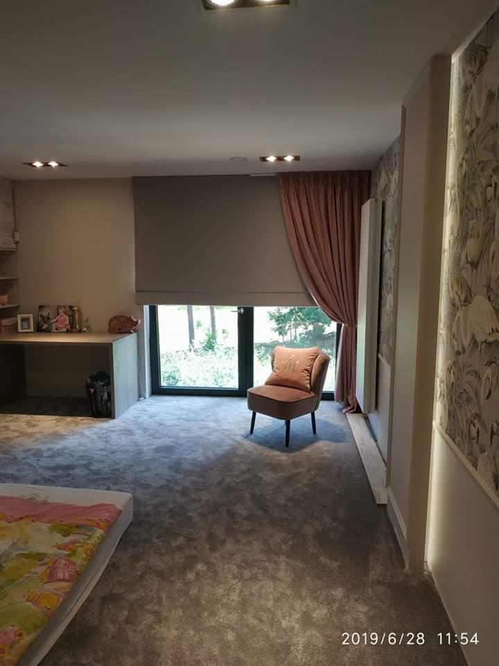 Pokój dziecięcy z szarym dywanem, różowym fotelem z monogramem, zasłoniętym oknem i tapetą w ptaki, oświetlony punktowymi lampami sufitowymi.