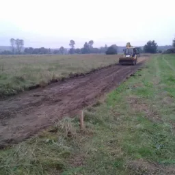 Żółta koparko-ładowarka CAT wyrównuje teren na łące, pozostawiając za sobą ślad opon w błocie; w tle drzewa i pochmurne niebo.