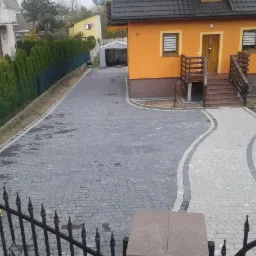 Ułożona nawierzchnia z kostki brukowej w dwóch odcieniach szarości, tworząca ścieżkę do domu i podjazd, widoczne ogrodzenie i żółta zagęszczarka w tle.