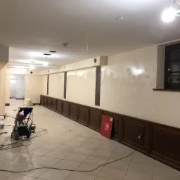 Wnętrze biura w trakcie malowania natryskowego, widoczny agregat malarski, ściany w odcieniach beżu, drewniana boazeria i plamy świeżej farby na ścianie pod oknem.