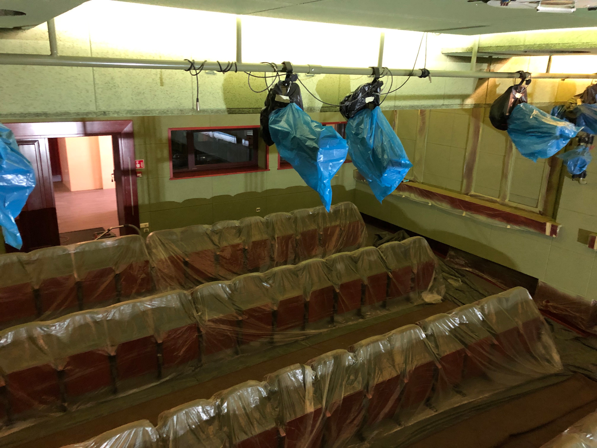 Sala kinowa w trakcie zabezpieczania przed pracami remontowymi: fotele zakryte folią ochronną, lampy wiszące zabezpieczone workami, ściany oklejone taśmą malarską.