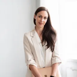 Kobieta w beżowej marynarce trzymająca laptopa w kolorze różowego złota, widoczny zegarek na ręce.