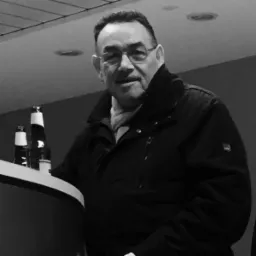 Czarno-białe zdjęcie mężczyzny w okularach i kurtce, stojącego przy barze z butelkami. Uśmiechnięty, z szalikiem, w pomieszczeniu z oświetleniem sufitowym.