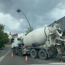 Ciężarówka betonomieszarki IFA z operatorem w żółtej koszulce na tle nowoczesnego budynku ze szklaną fasadą i ulicy z samochodami, oświetlona latarnią uliczną.