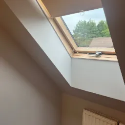 Pomalowane wnętrze poddasza z oknem dachowym z drewnianą ramą, widok na zieleń drzew za oknem, biały grzejnik pod oknem, gładkie ściany w odcieniach beżu i bieli.