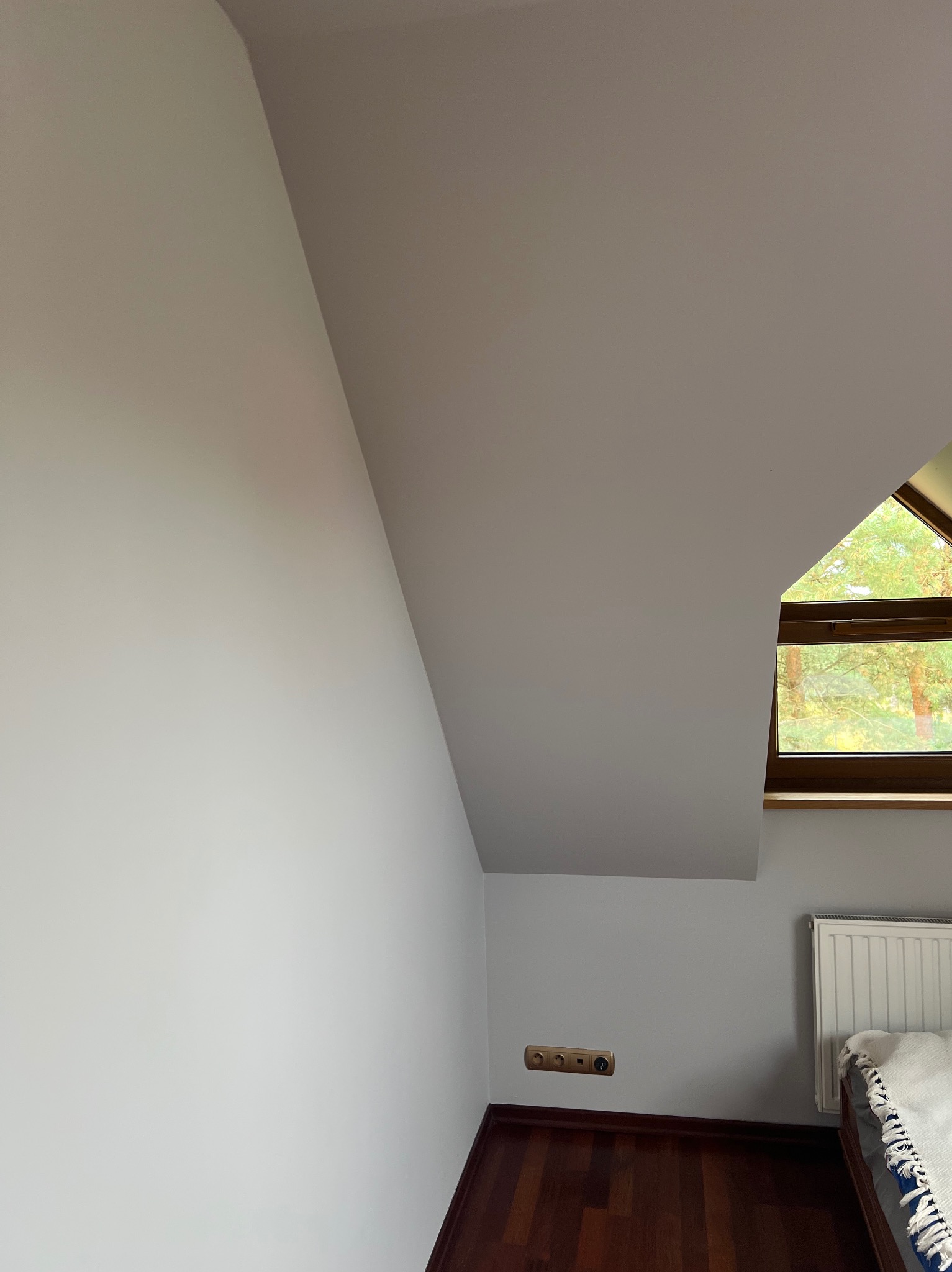 Pomalowane na biało poddasze ze skośną ścianą, oknem dachowym i grzejnikiem, widoczne gniazdka elektryczne i fragment łóżka.