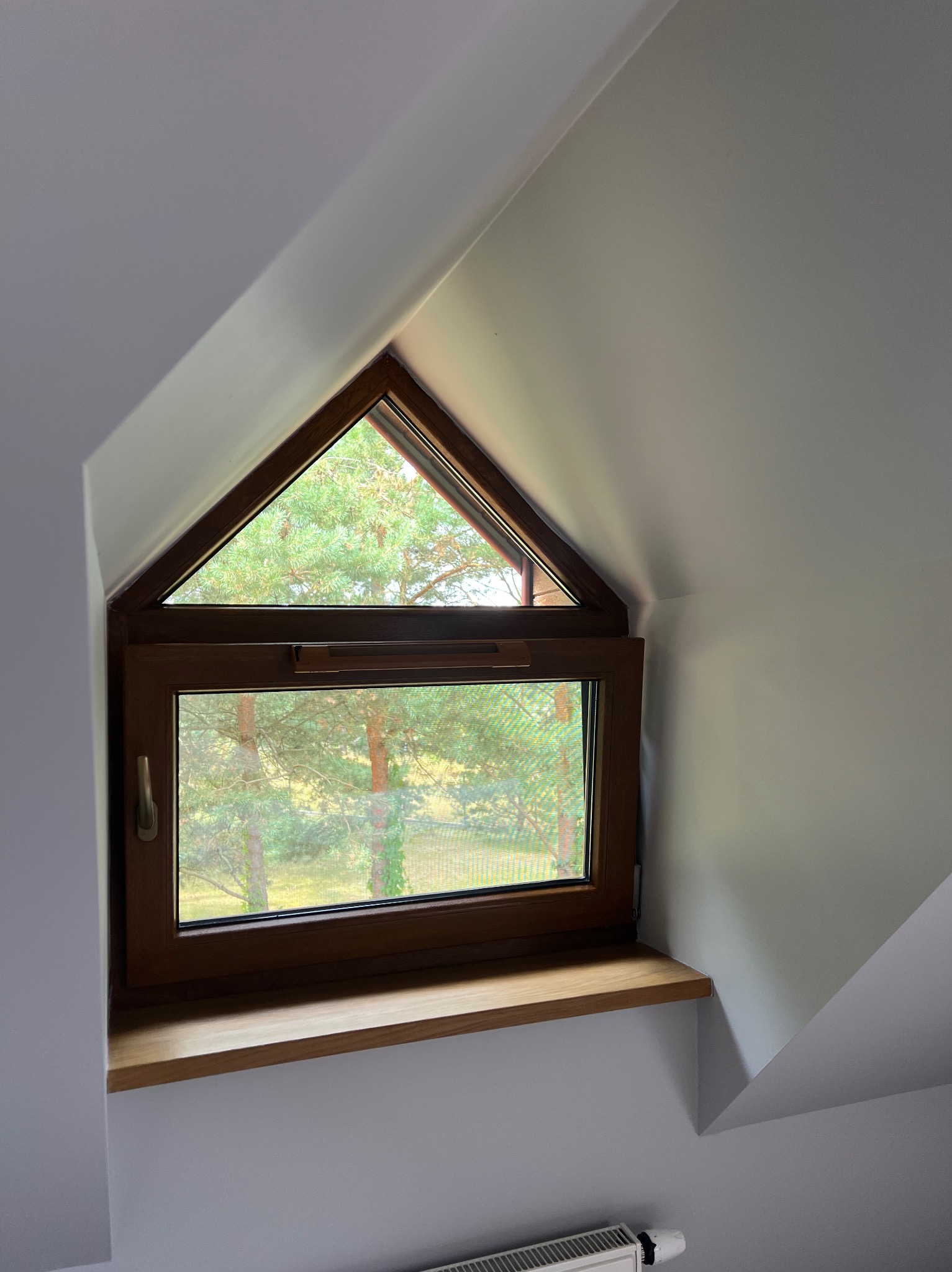 Trójkątne okno na poddaszu z drewnianą ramą i parapetem, widok na zewnątrz na drzewa iglaste i siatkę ogrodzeniową.