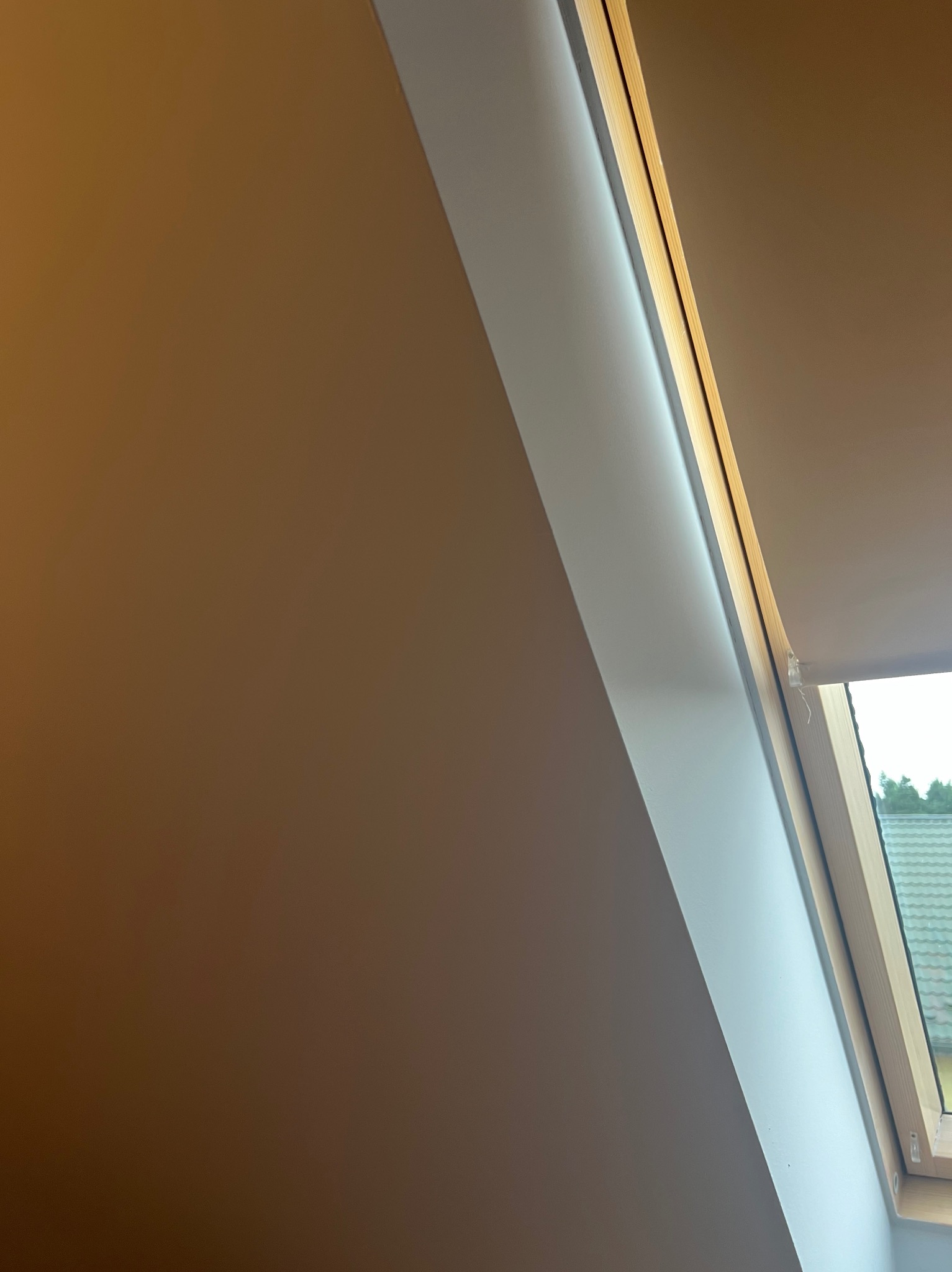 Pomalowane na brązowo poddasze ze skośną ścianą i oknem dachowym z drewnianą ramą, fragment widoku za oknem na zewnątrz.