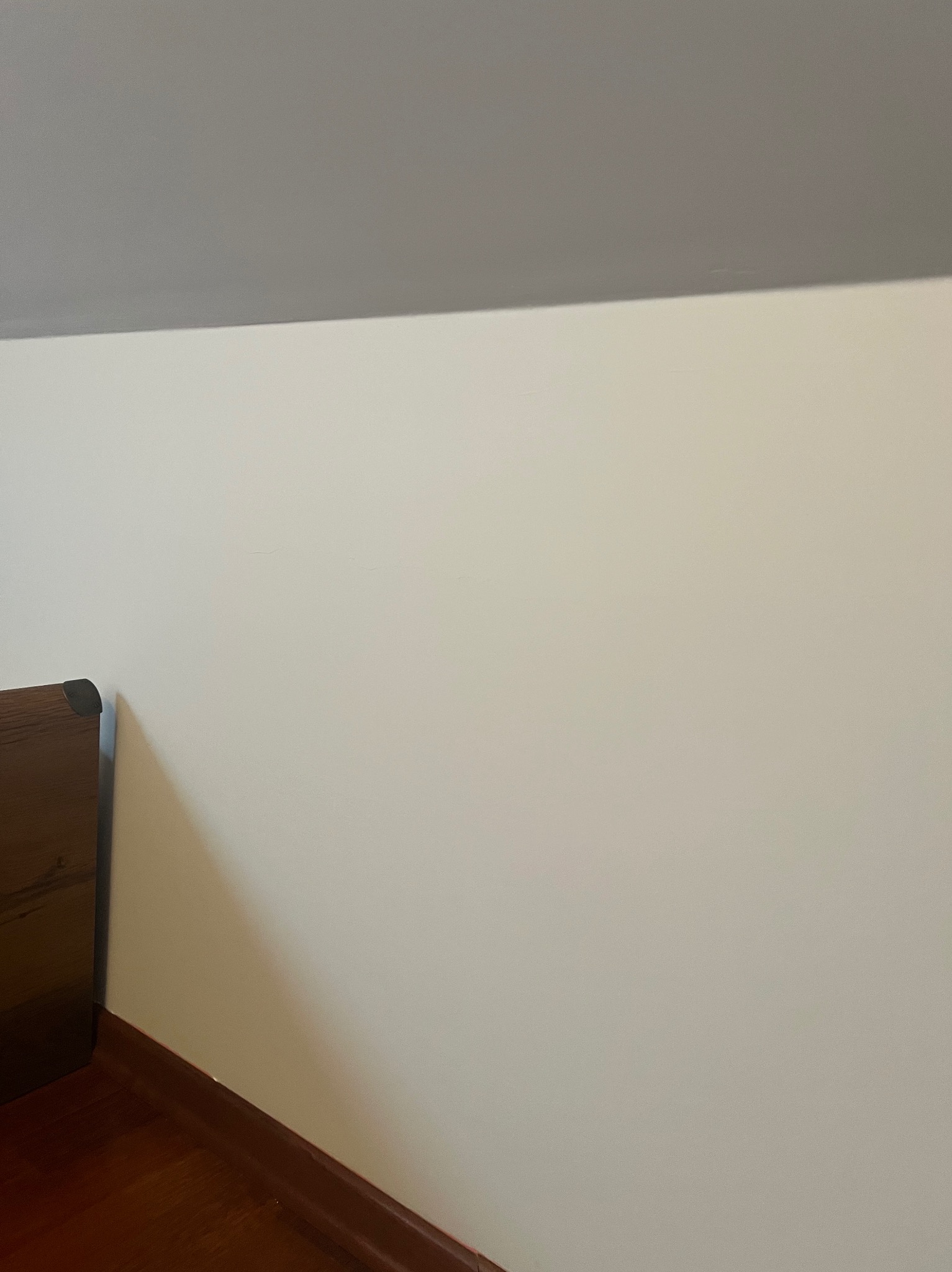 Fragment ściany pomalowanej na biało, z widocznym fragmentem drewnianej ramy i podłogi z brązowymi listwami przypodłogowymi, górna część kadru z szarym sufitem pod skosem.