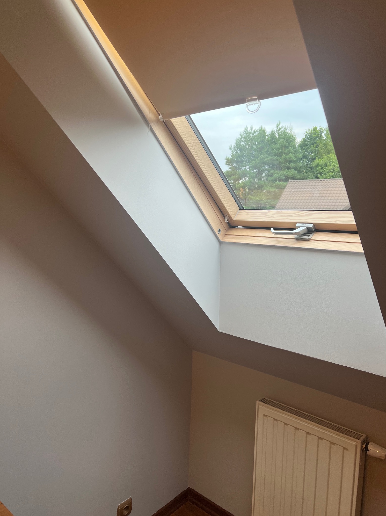 Pomalowane wnętrze poddasza z oknem dachowym z drewnianą ramą, widok na zieleń drzew za oknem, biały grzejnik pod oknem, gładkie ściany w odcieniach beżu i bieli.