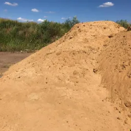 Duża pryzma sypkiego, żółtego materiału z widocznymi grudkami i nierównościami, składowana na placu budowy, w tle trawiasta skarpa i błękitne niebo z chmurami.