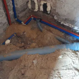 Instalacja wodno-kanalizacyjna w piasku, widoczne rury PCV szare i rury PEX czerwono-niebieskie w surowym wnętrzu budynku. Fragment instalacji w trakcie montażu.